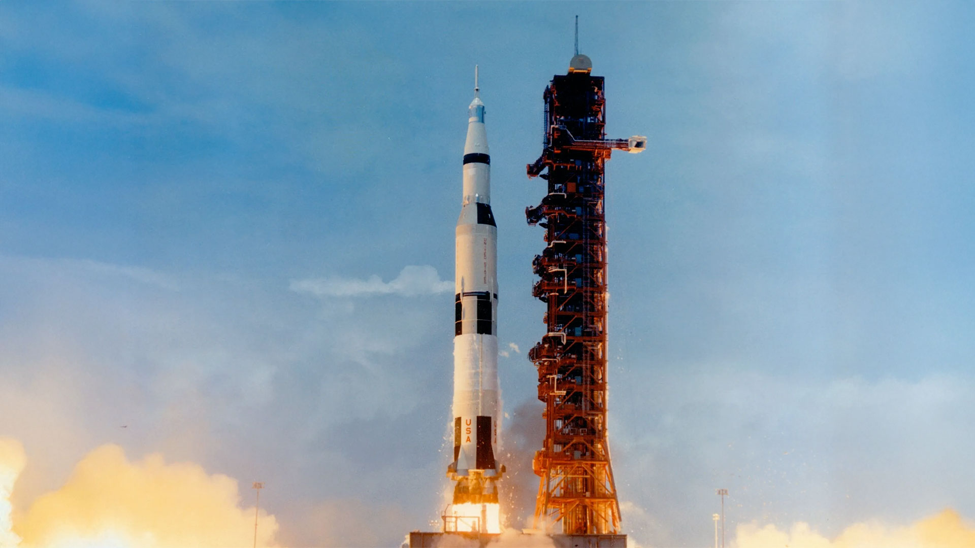 a white rocket with black stripes lifts off alongside a launch support tower into the blue sky