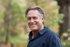 A casually-dressed man, appearing to be in his 50s, smiles at the camera. His hair is a bit long and messy. He is handsome and relaxed.