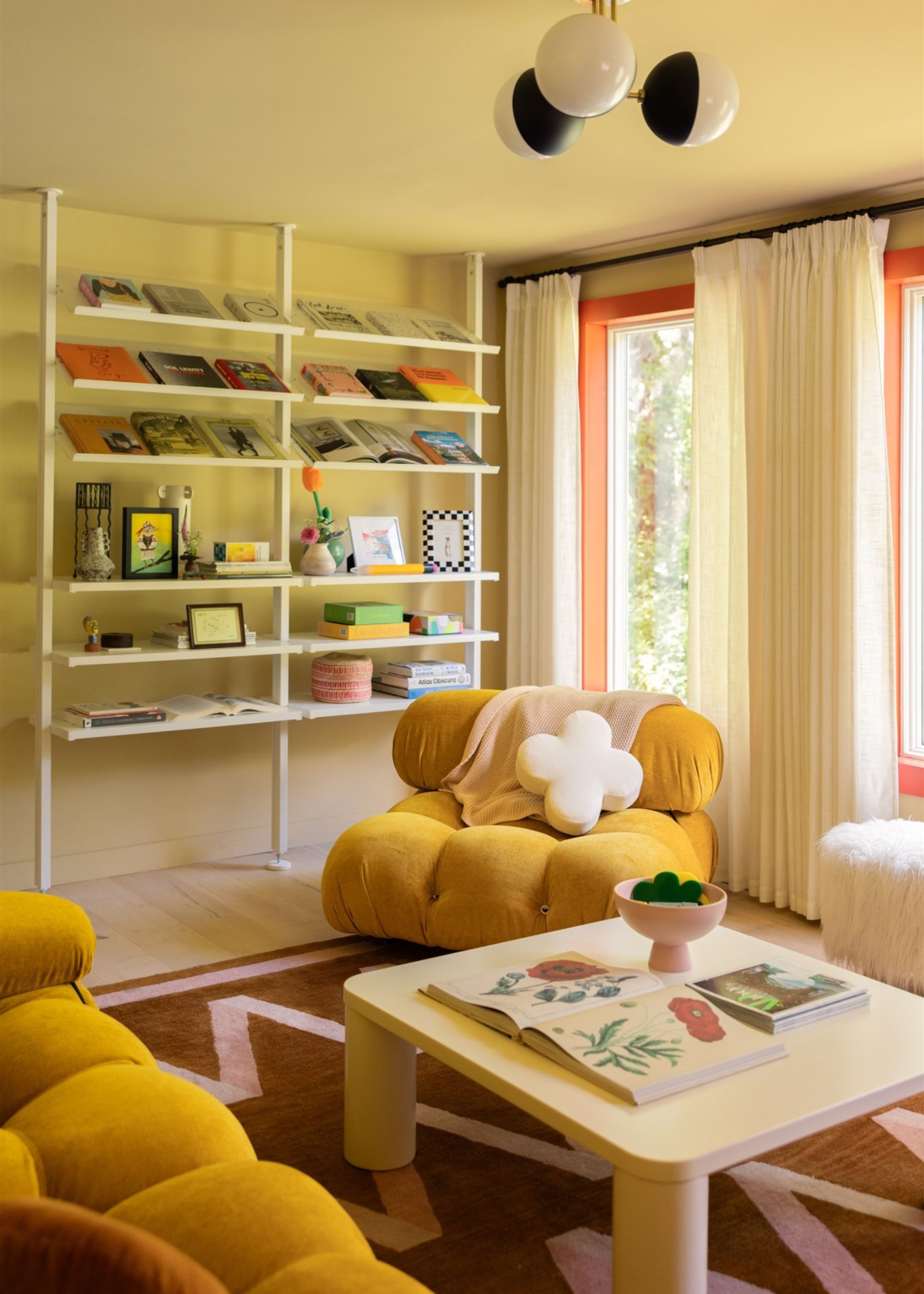 A playful living room with butter-yellow color-drenched walls, large windows with tomato-red trim, sheer curtains, and a wall of white open shelving.