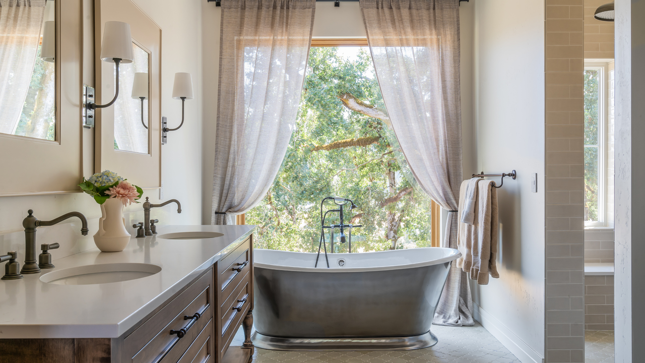 A bath tub and a large window with curtain draping&#039;s