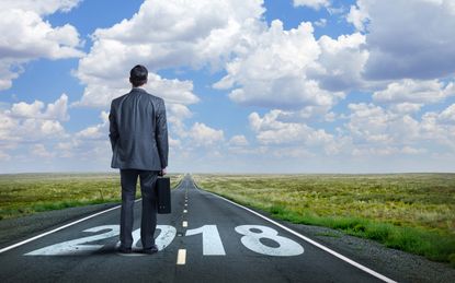 A businessman stands while holding his briefcase as he looks down a long, straight, rural road that has "2108" painted on it.In the distance, as the road meets the horizon, puffy clouds punct