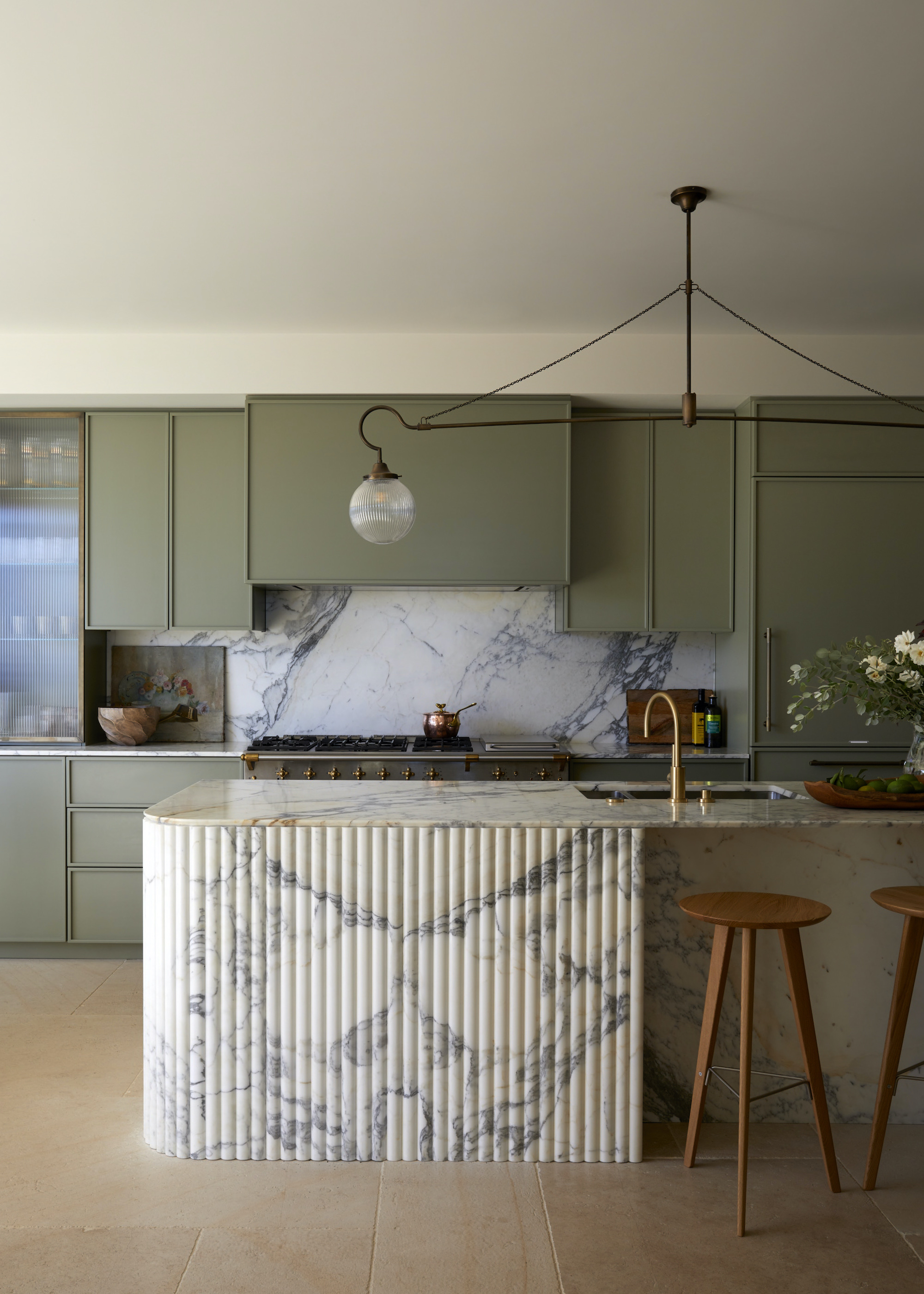 a sage green kitchen with fluted marble island, wooden stool and a large pendant across the space