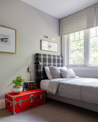 gray boy's bedroom with upholstered black and gray check headboard, grey patterned blind and bedding, red trunk, black wall light, artwork