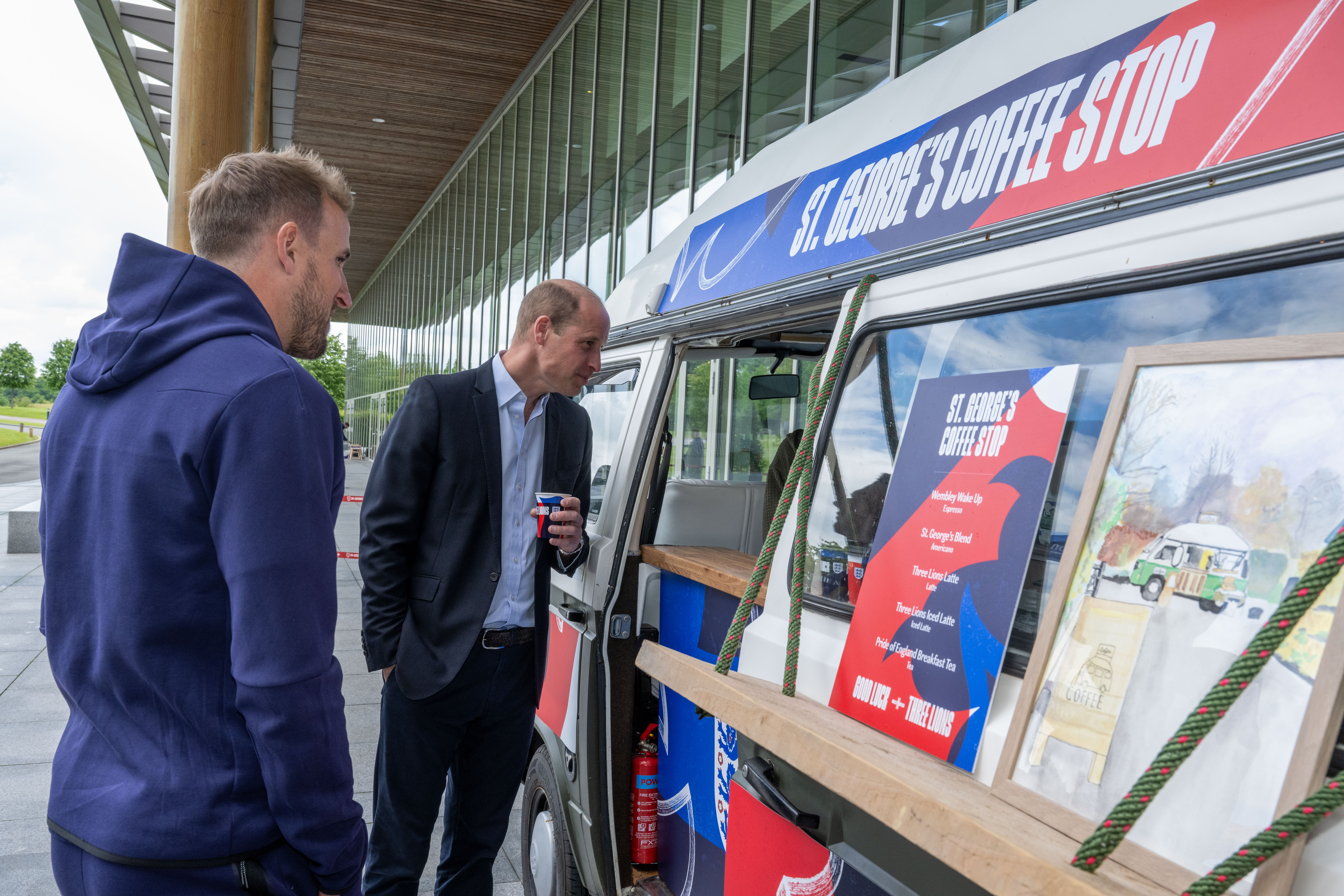 Prince William holding a coffee talking to someone in the window of a coffee stand