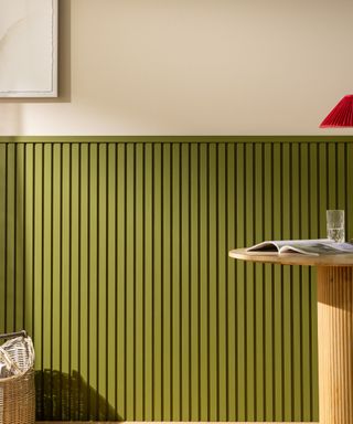 A green wooden wall panel with a woven storage basket to the left, a curved wooden table with a magazine, glass, and red lamp on to the right, and a beige wall strip with a framed white wall art print above it