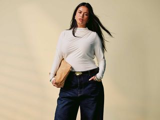 model wears long-sleeve white blouse, dark wash baggy jeans, and a tan clutch while posing for a picture and hair blows in the wind.