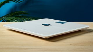 a white smart scale by withings rests on a wooden table, showing its LED screen and weighing scales