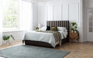 white bedroom with white painted paneling on walls, herringbone floor, duck egg blue rug, upholstered bed with white bedding patterned blanket and cushions, wooden side table, table lamps