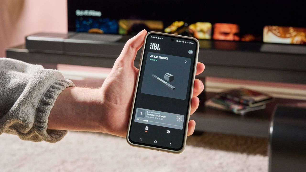 Man holding phone in front of JBL Bar 1300MK2 with JBL One app displayed