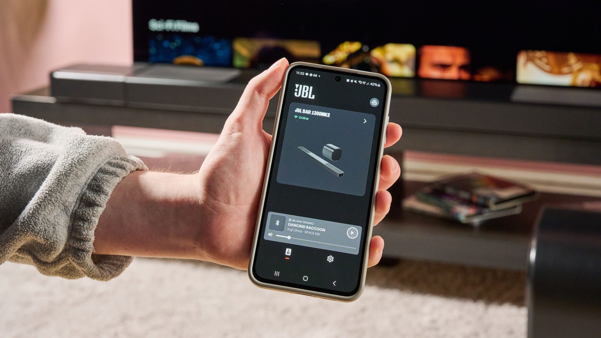 Man holding phone in front of JBL Bar 1300MK2 with JBL One app displayed
