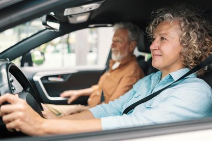A middle-aged woman drives a car, with a senior man in the passenger seat. 