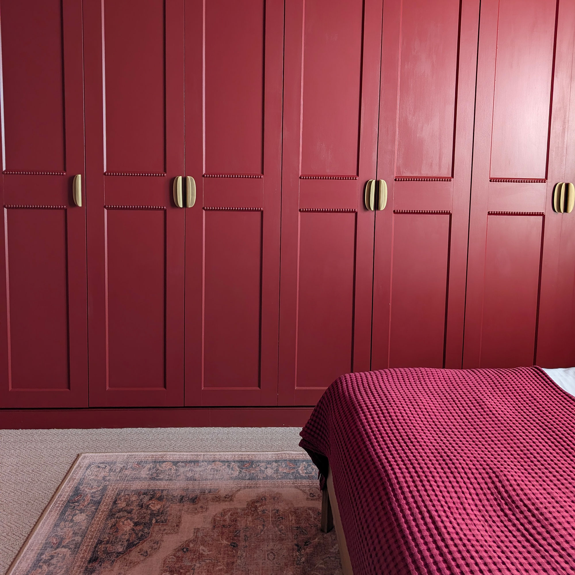 Bedroom with red panelled built in wardrobes with brass pulls, red throw on bed, neutral carpet and pink and blue faded Persian style rug on floor