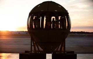 a metal orb-shaped capsule lined with windows in a hangar