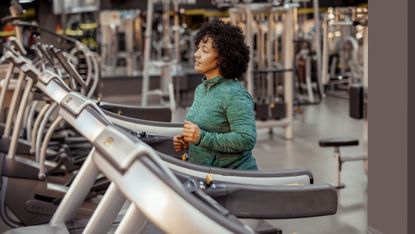 A woman in a green hoodie walks on a treadmill in a gym setting. She is smiling slightly and has her arms at waist-height 