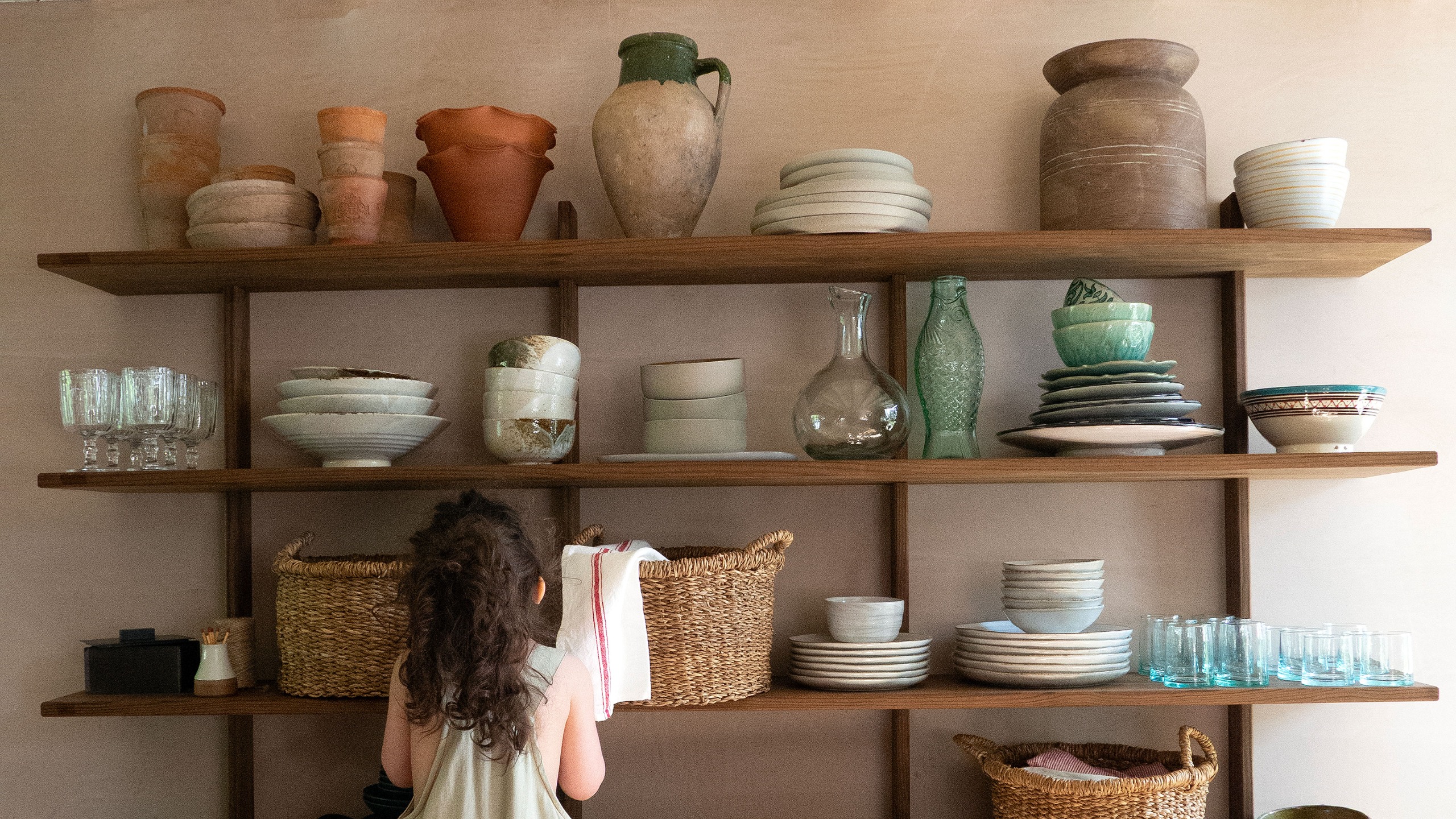 Small girl standing in front of open wooden shelving filled with ceramics, glassware, jugs and baskets