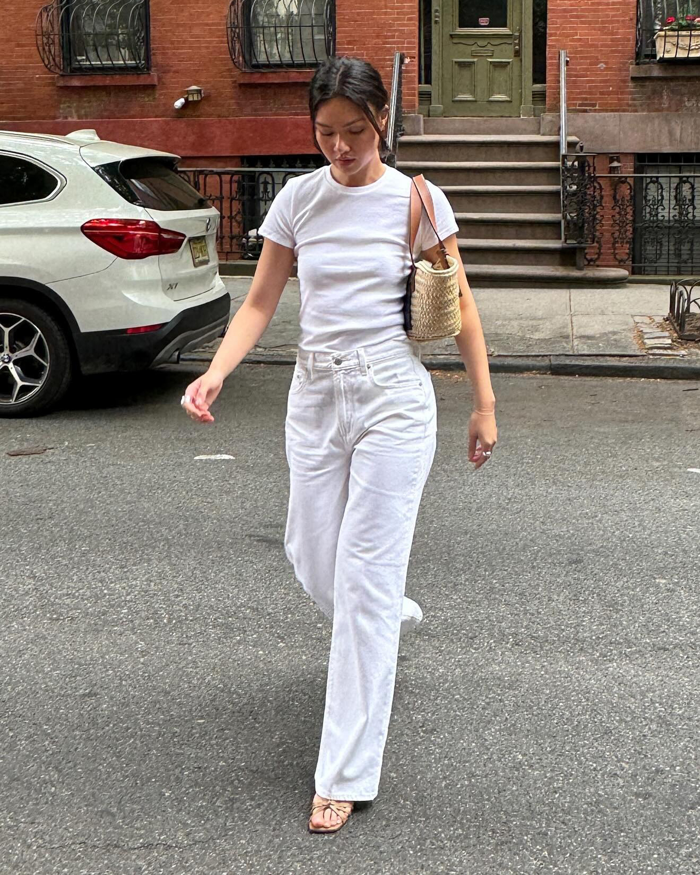 style influencer Mimi Nguyen walks across a New York City street wearing a small straw tote bag with brown leather straps, a fitted white T-shirt, white high-waisted straight-leg jeans, and neutral strappy heeled sandals