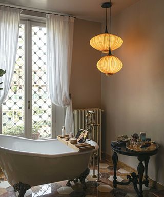 A bathroom with white sheer drapery, a standalone bathtub, and a bespoke glowing light fixture hanging above