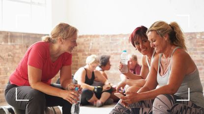 Three women sitting together in light and airy gym after a cardio vs strength training workout class, drinking from water bottles and talking