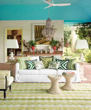 spring living room, outdoor, with turquoise blue ceiling, shiplap, white couch with green patterned pillows, armchair, beaded stools, large table lamps, console table with candlesticks, artwork, beaded chandelier, terracotta tiled flooring