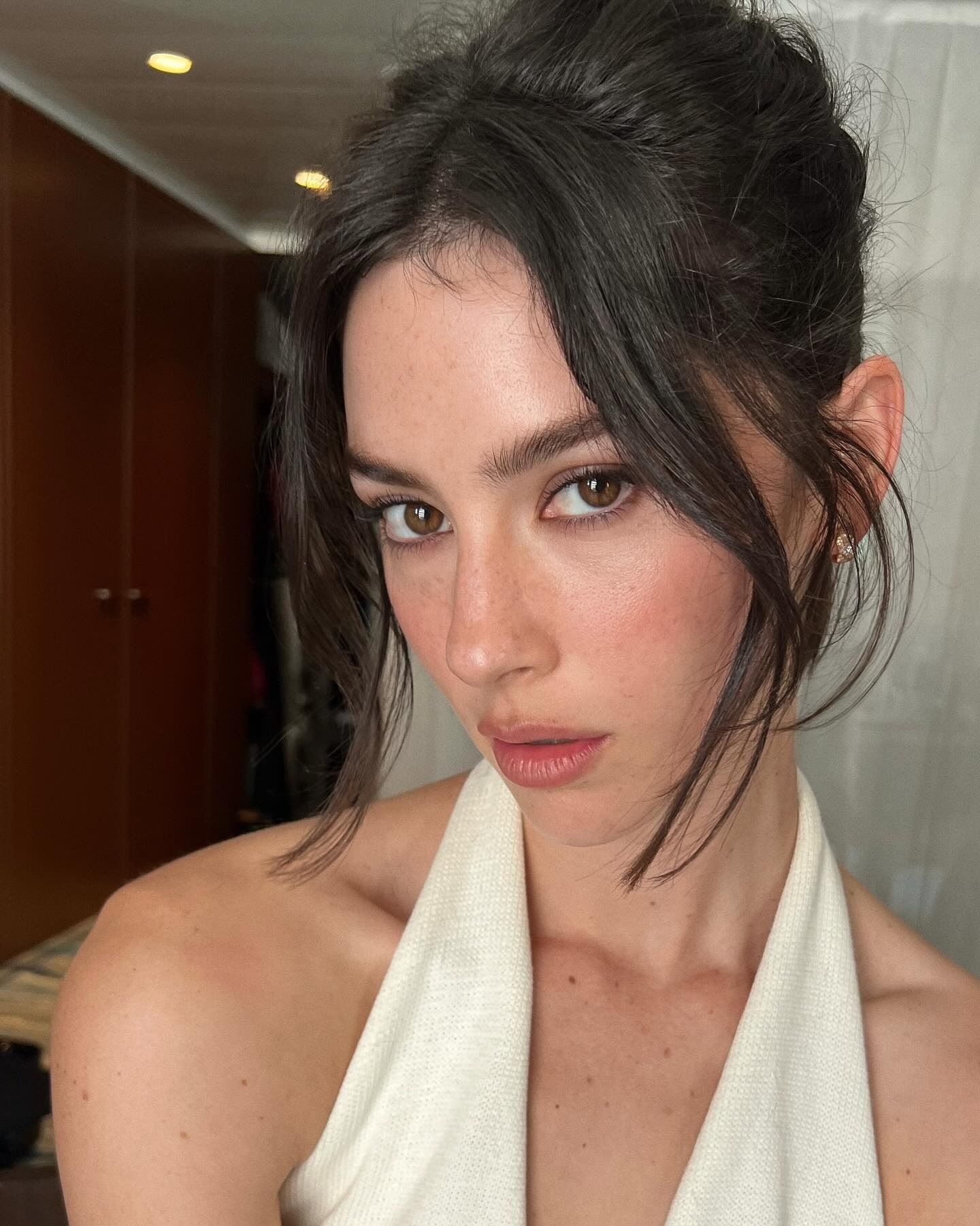 Gracie Abrams with lightweight makeup and an updo with face-framing tendrils wearing a white halterneck dress