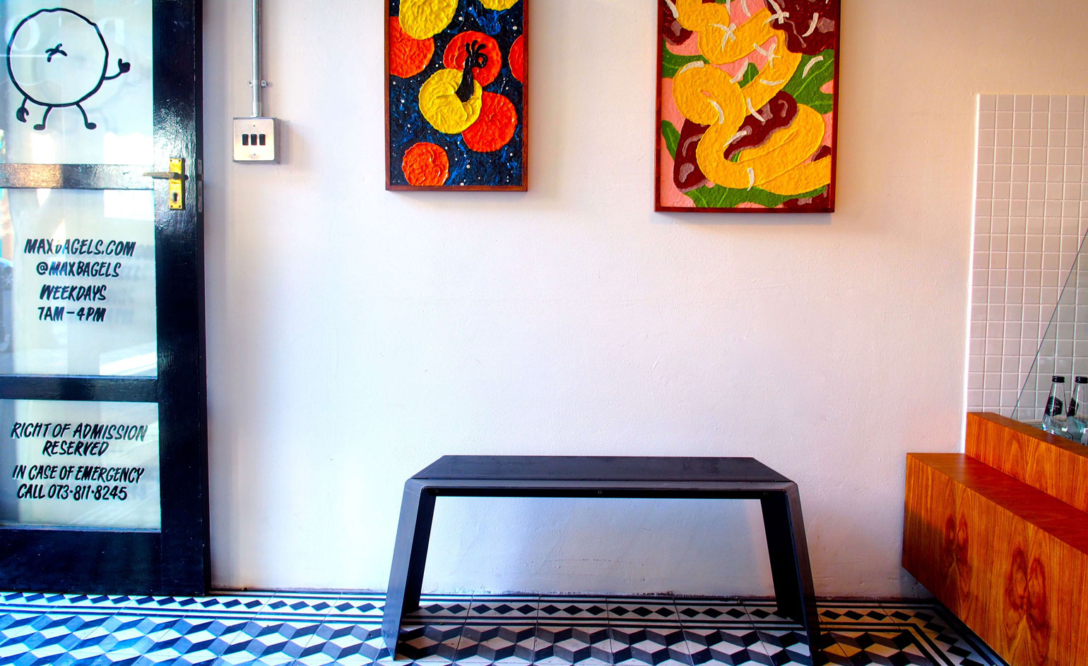 Alternative view of Max Bagels featuring light coloured walls, a black and glass door with shop information, colourful wall art, a black bench style seat, geometric flooring and a partial view of a wooden counter