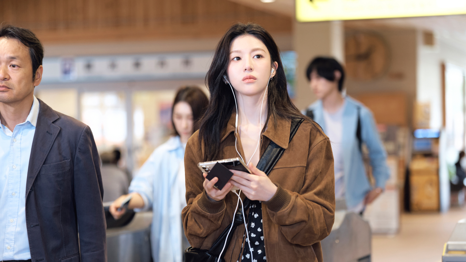 A woman (Go Youn-jung as Cha Mu-hee / Do Ra-mi) wears wired headphones and holds a foldable smartphone as she stands in a lightly crowded train station. A still from the K-drama 'Can This Love Be Translated?'