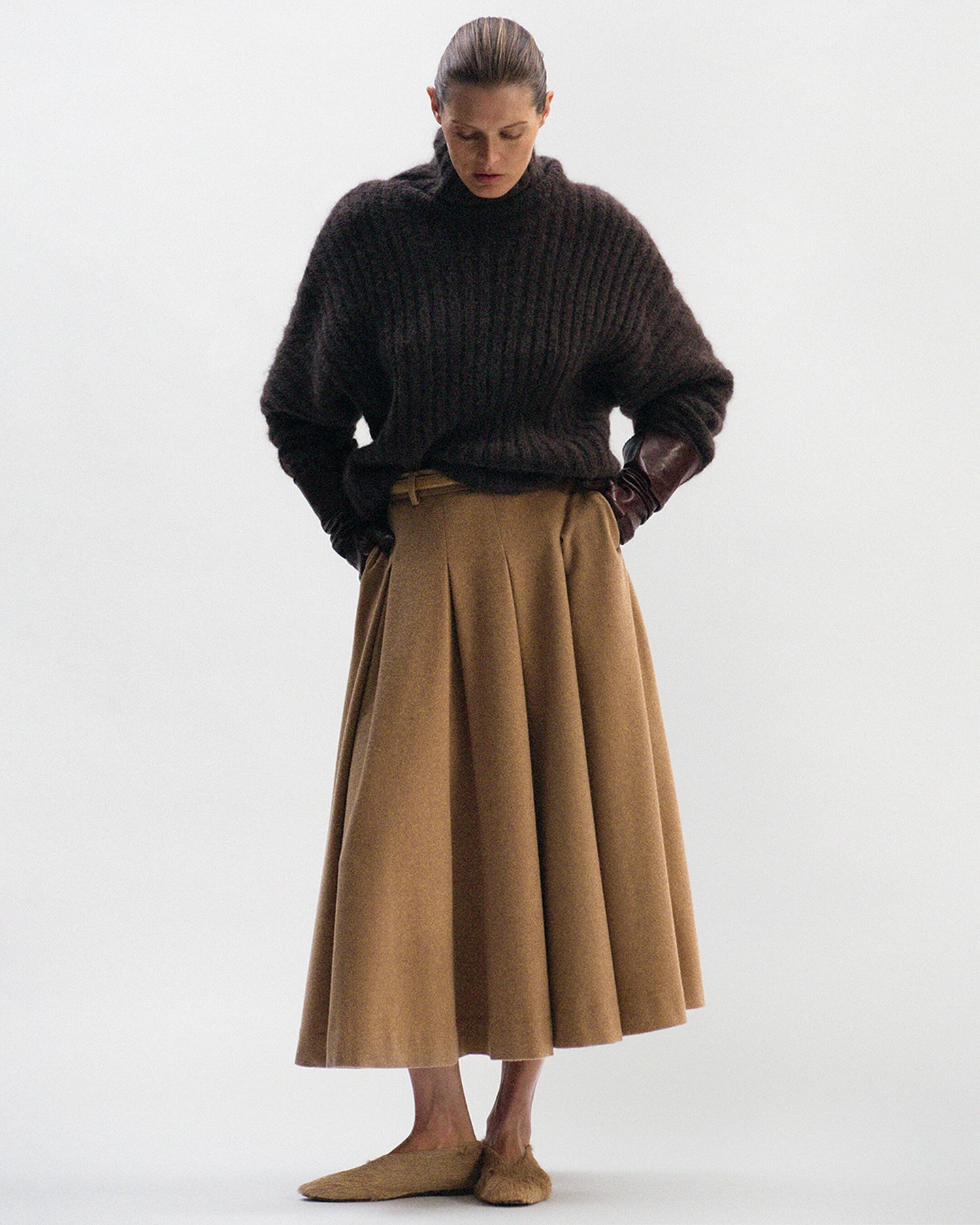 A woman wears a brown sweater, tan wool skirt, and tan flats.