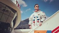 a man wearing a white spacesuit and sun glasses stands on a mobile stairway outside a 60's style airport terminal