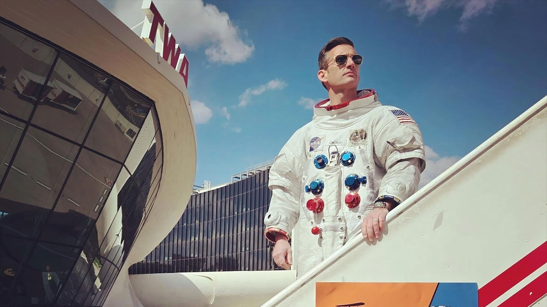 a man wearing a white spacesuit and sun glasses stands on a mobile stairway outside a 60's style airport terminal