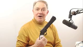 Dennis Bateman, the voice actor for TF2's Spy, holds up a prop revolver while smiling at the camera.