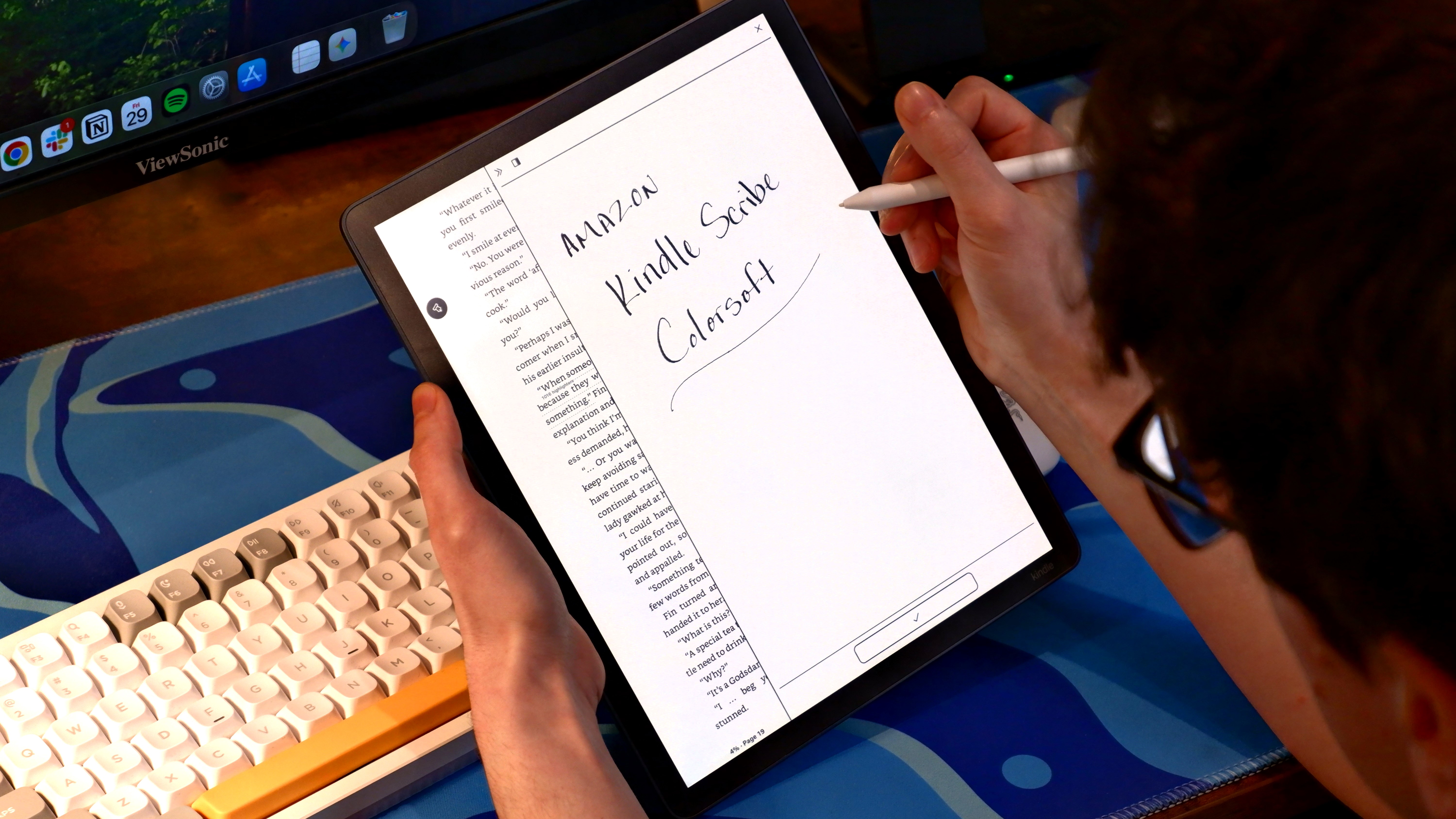 Amazon Kindle Scribe Colorsoft held over a desk with a pen in hand, writing "Amazon Kindle Scribe Colorsoft."