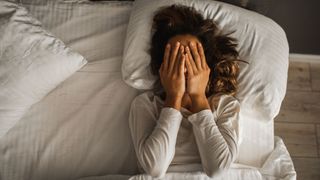 A woman with brown hair lies in bed with her hands over her face after having a nightmare