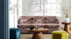 Floral-patterned sleeper sofa styled in a sunlit living room alongside wooden tables, a sculptural floor lamp, and jewel-toned ottomans