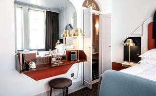 Interior view of a room at Henrietta Hotel, London, UK featuring patterned flooring, dual coloured walls, brass lamps, a wall-mounted mirror and a stool under a wooden shelf with books, a phone, glasses and bottles of drinks on top. The bed, window and curtains can be seen in the reflection of the mirror