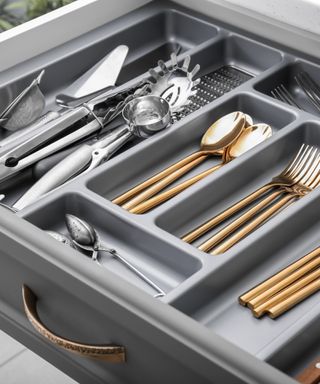 An image of a gray wood kitchen drawer with a gold accent handle that's open with a gray plastic utensil caddy filled with a mix of silver and gold cutterly and utensils