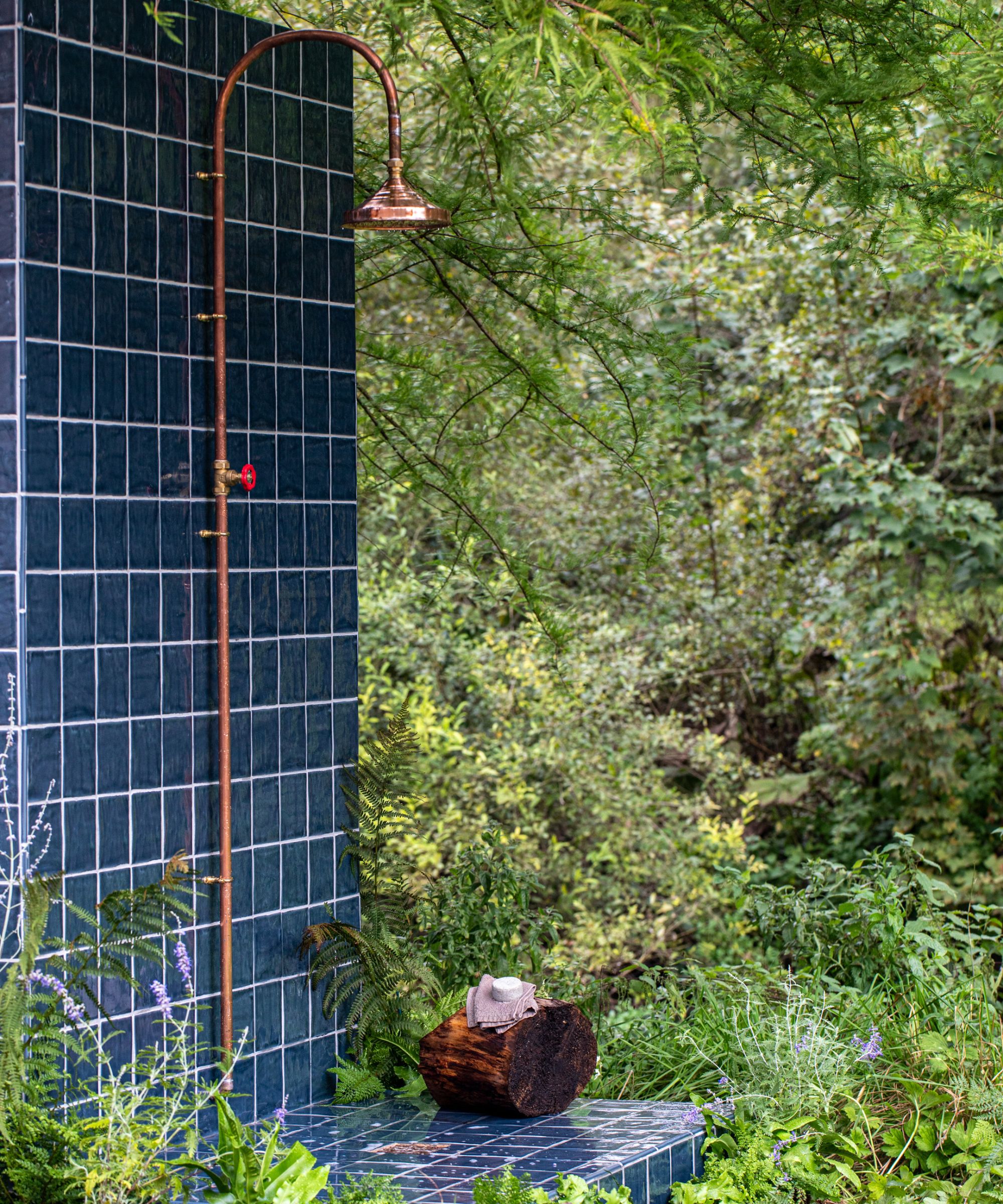 A dark navy square-tiled outdoor shower wall with a copper rainfall head and red-handled tap, surrounded by lush ferns and woodland planting.