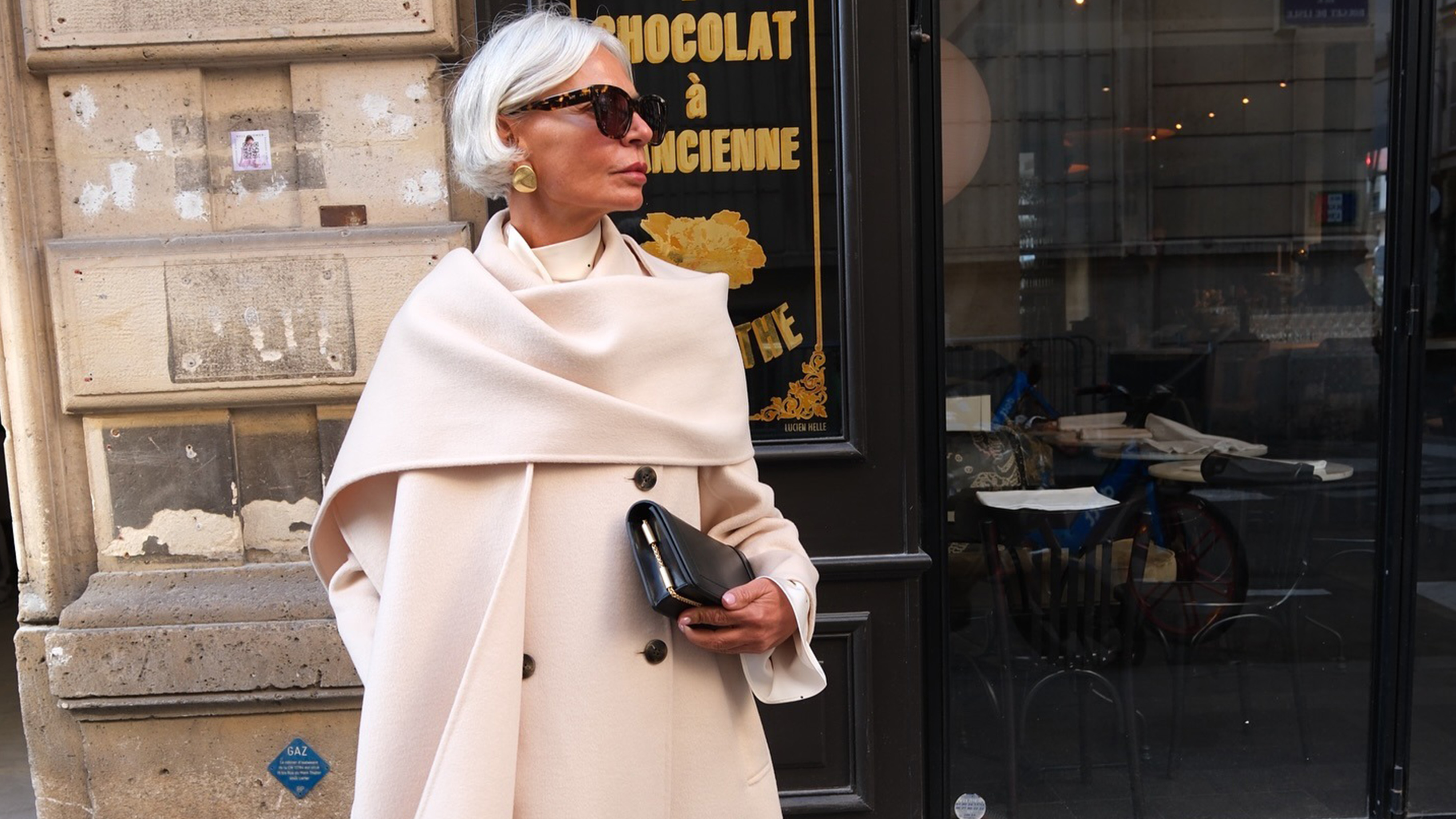 Grece wears an ivory wool coat with an attached scarf and a black clutch.
