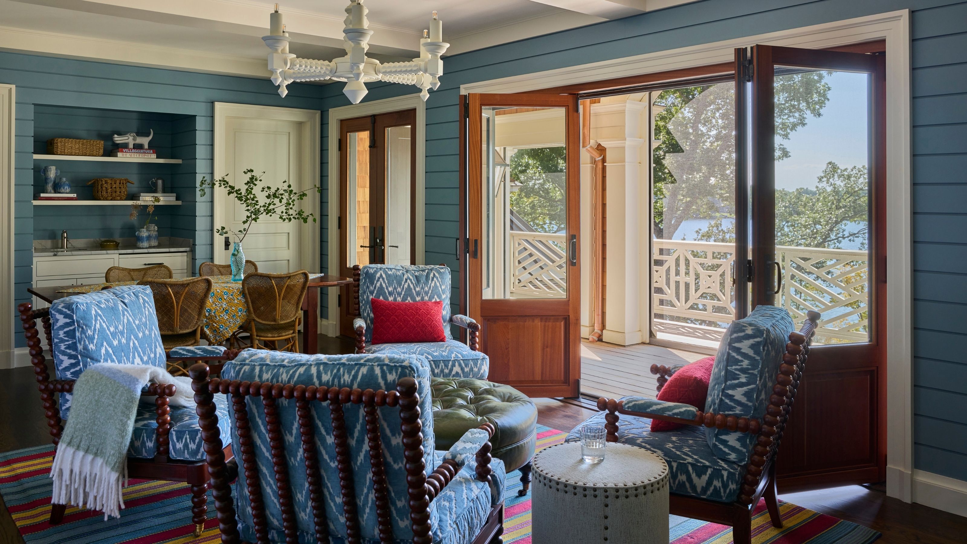 Blue shiplap living room with patterned armchair seating surrounding a green ottoman, highlighted by an open folding glass door that leads to a sunny balcony overlooking water views.