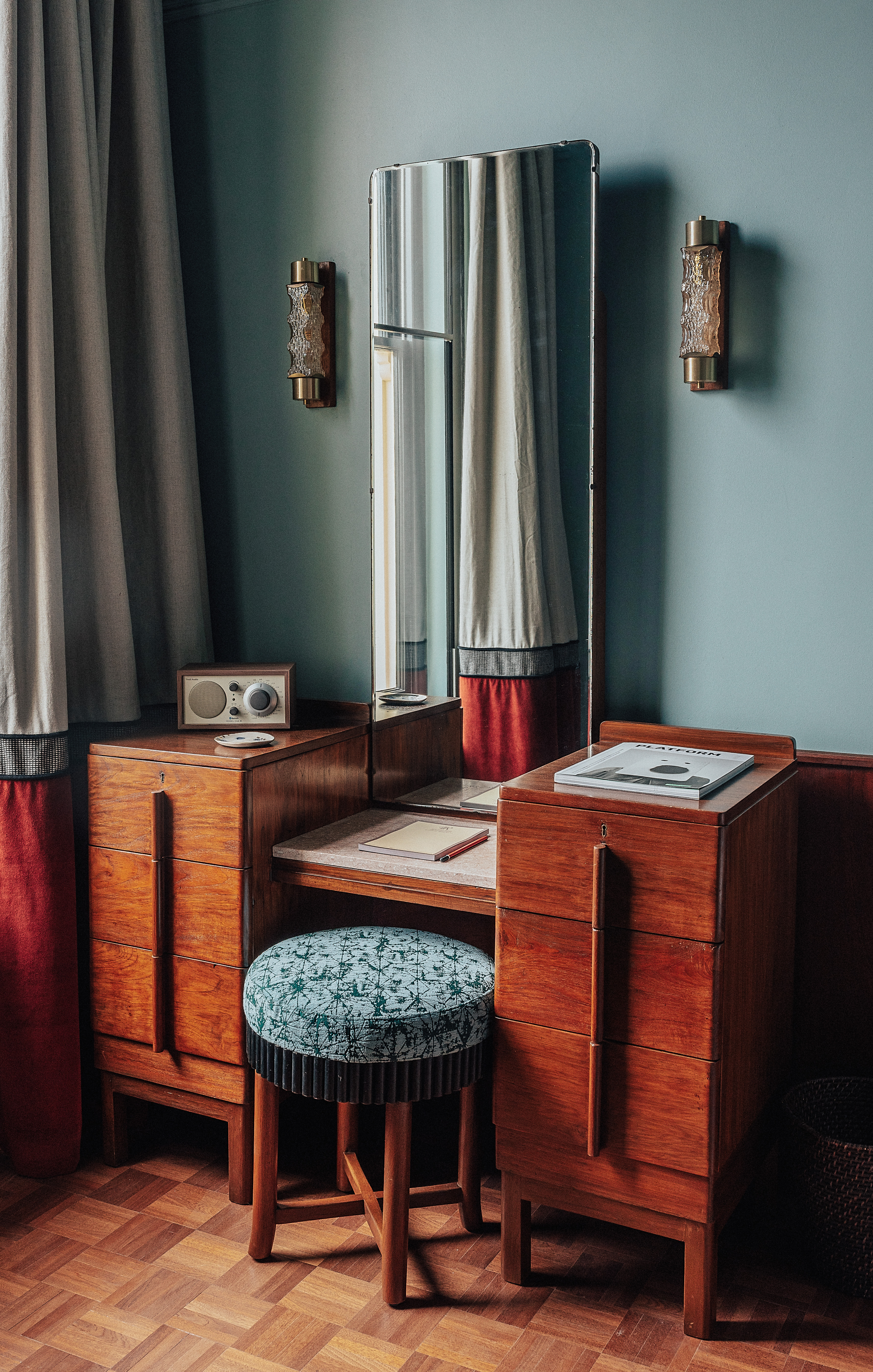 A vibrantly decorated hotel suite inspired by Bombay residences features buttery or tile blue walls adorned with original portraiture artworks, bespoke retro-fueled furnishings in wood, leather, and patterned fabric, and thriving plants.