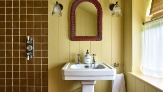 A yellow color-drenched bathroom with wall panelling, a white sink, an arched dark brown mirror, and two wall sconces. Caramel square wall tiles in the shower and a caf&eacute; curtain on the window. 