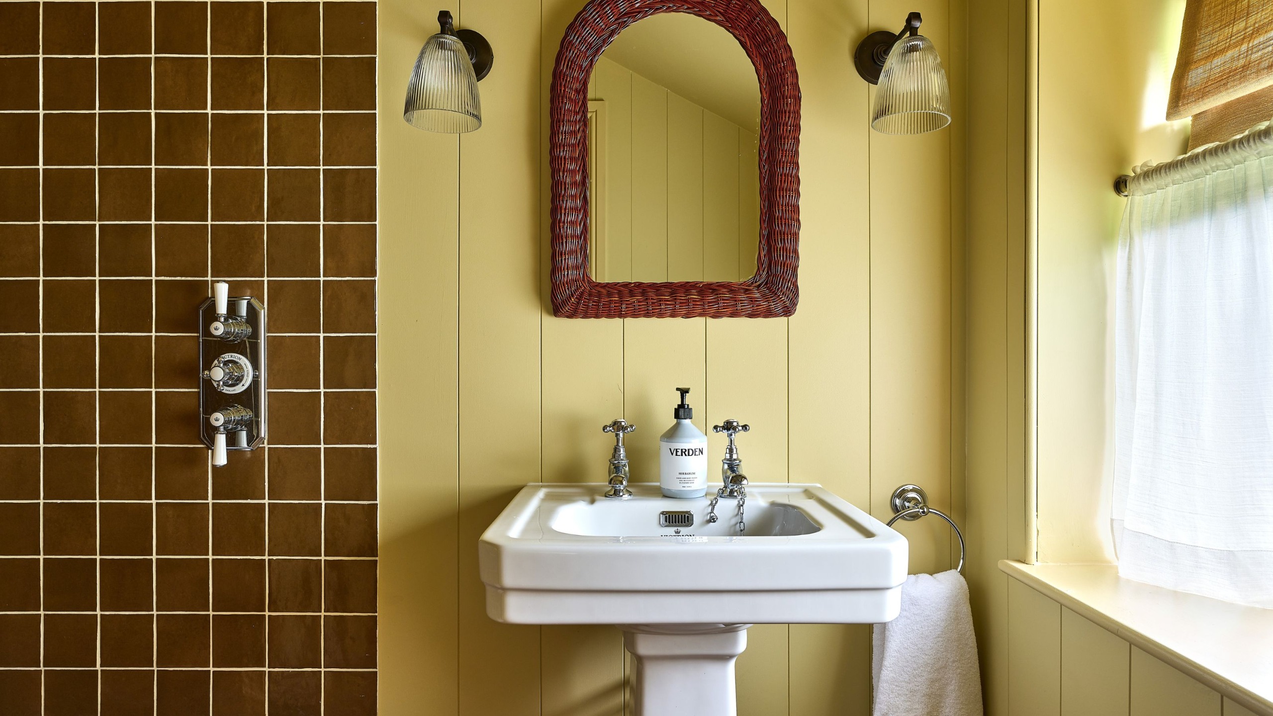 A yellow color-drenched bathroom with wall panelling, a white sink, an arched dark brown mirror, and two wall sconces. Caramel square wall tiles in the shower and a caf&eacute; curtain on the window. 