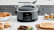 A Ninja PossibleCooker Pro Multicooker in a kitchen next to spaghetti and meatballs, blondies, slides, steak and a flatbread
