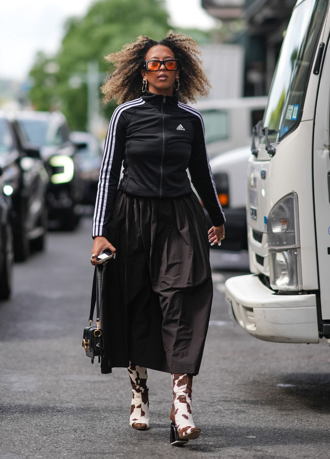 A guest wears black rimmed sunglasses with orange lenses, silver hoop earrings, black Adidas tracksuit jacket, black ruffled midi skirt, gold rings, white and dark brown cow pattern/print heeled boots, black leather bag, outside Tibi, during the New York Fashion week Spring/Summer 2025 on September 07, 2024 in New York, New York