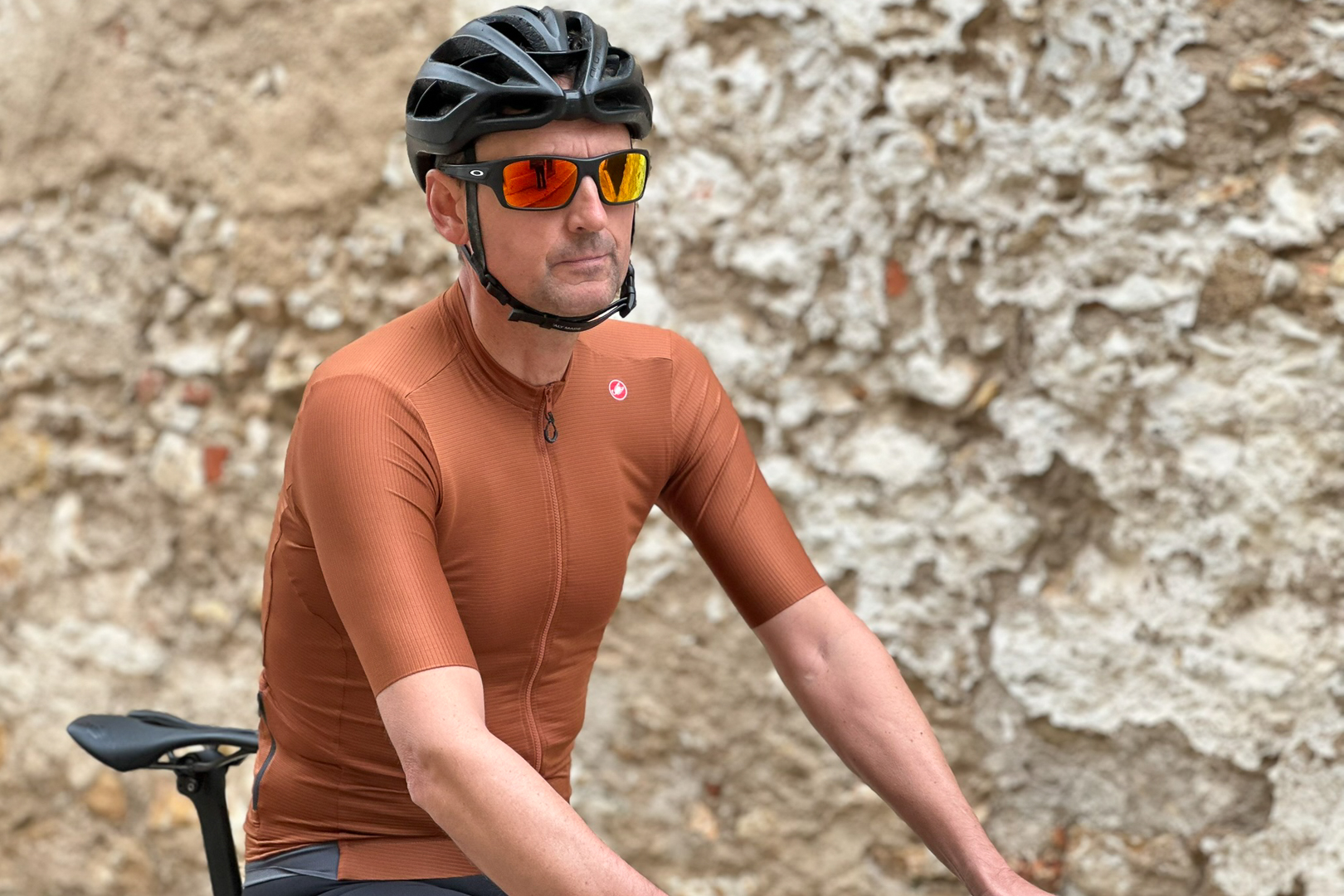 Man wearing a black helmet and brown cycling jersey standing near an old brick wall holding a bike