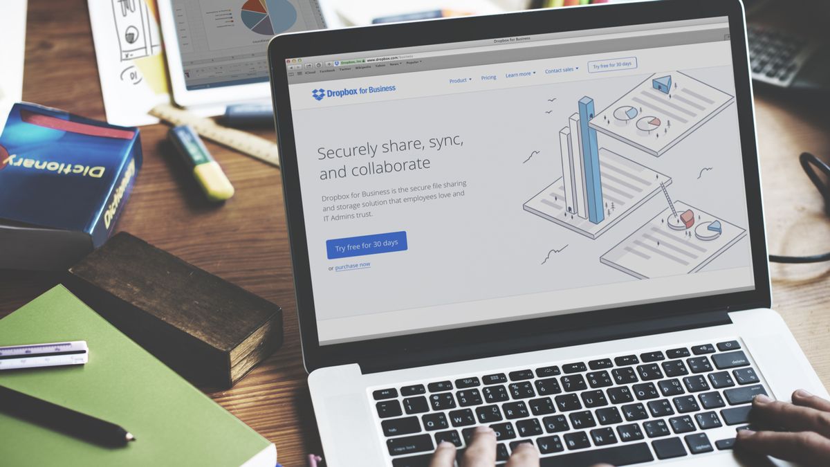 A MacBook on a cluttered desk displaying the Dropbox web page