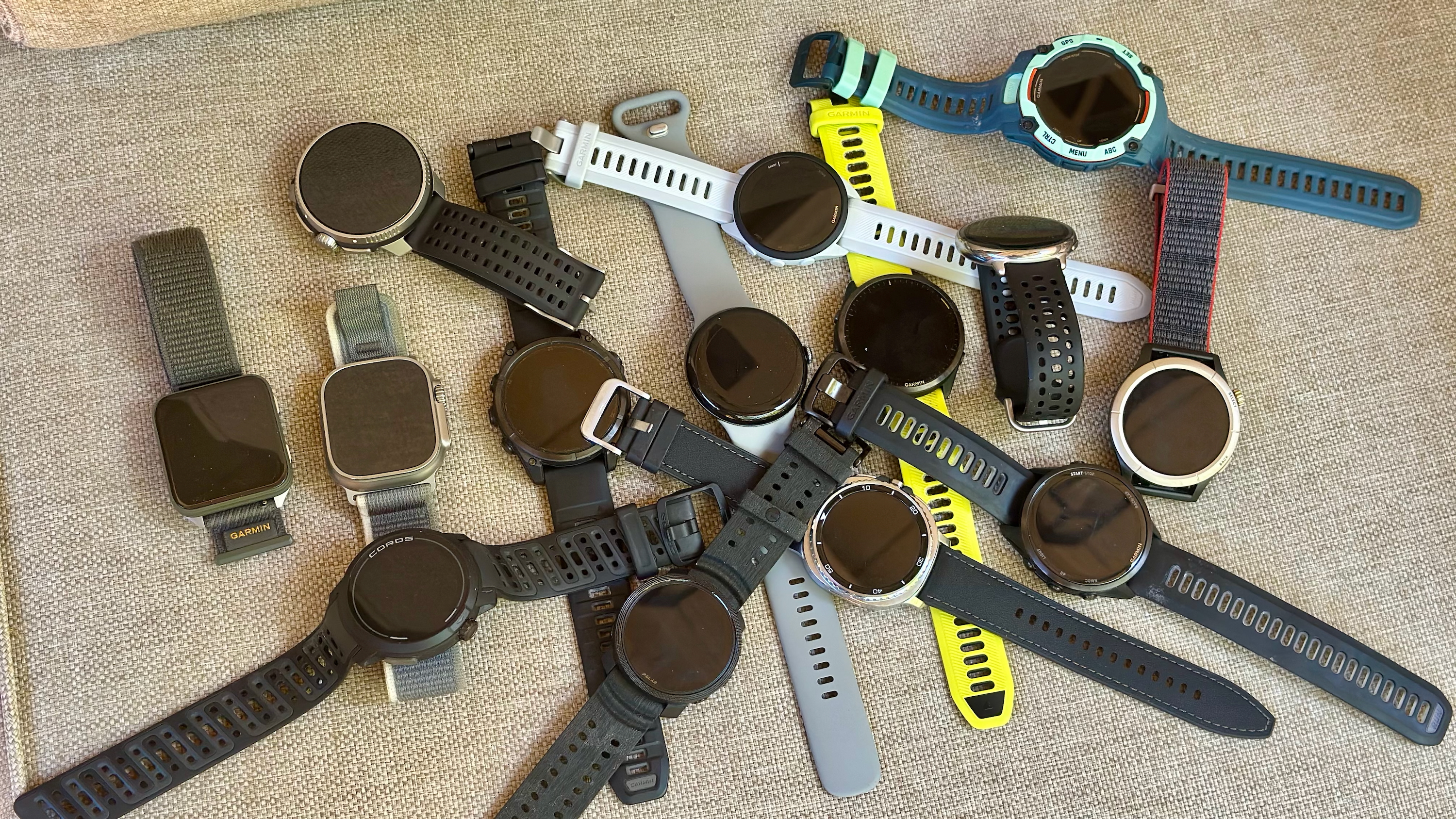 14 running watches and smartwatches sitting in a heap on top of a chair cushion, from brands like Garmin, COROS, Polar, Amazfit, Apple, Google, and Samsung.