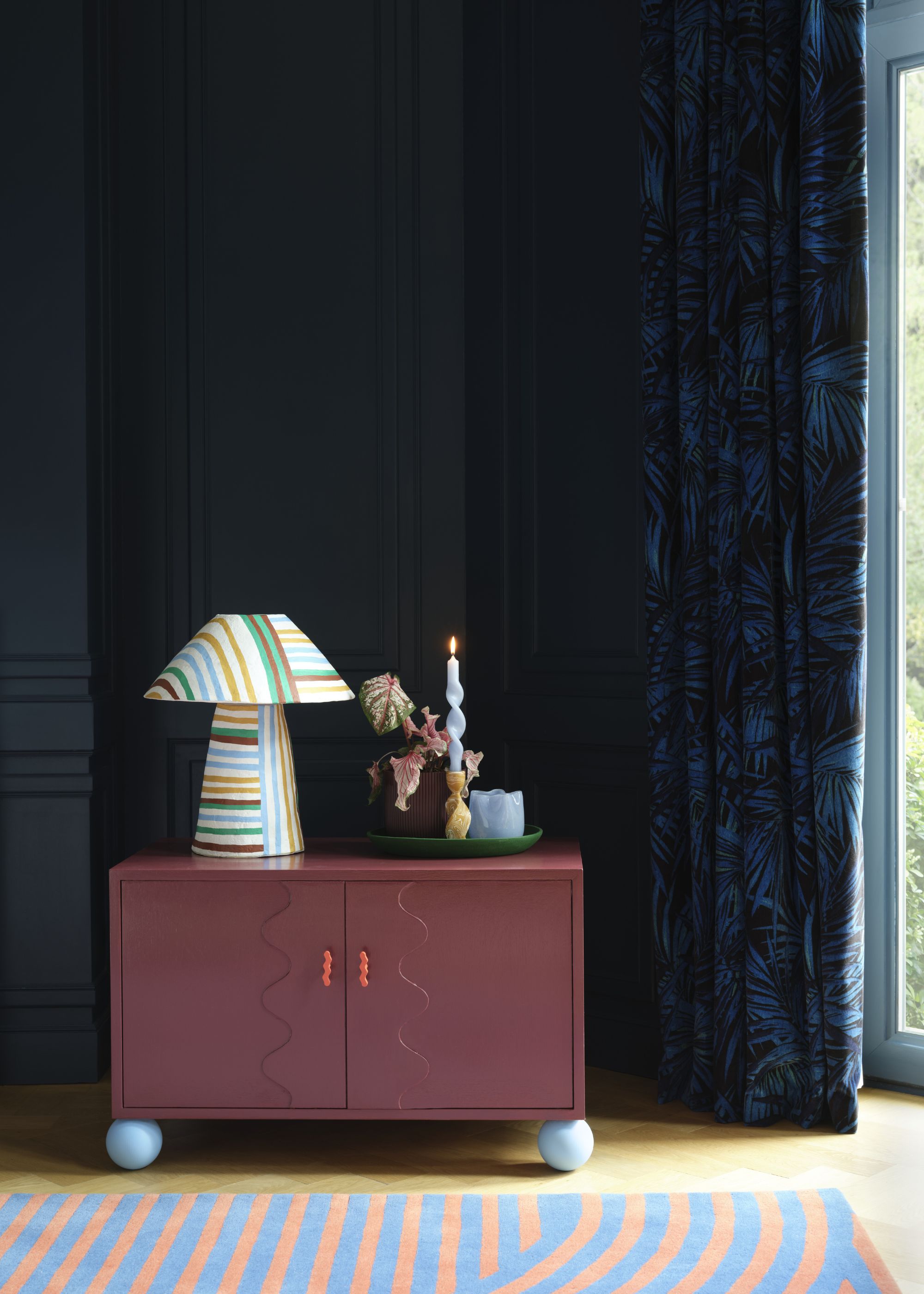 Corner of living room with dark blue walls and curtains, a blue and orange striped rug, and a burgundy dresser.
