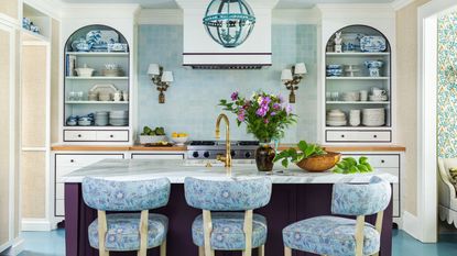 Blue and white kitchen with blue tiled backsplash, built-in alcoves with open shelves of china, aubergine painted island and bar stools upholstered in blue floral fabric