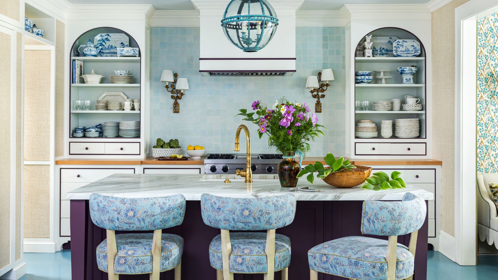 Blue and white kitchen with blue tiled backsplash, built-in alcoves with open shelves of china, aubergine painted island and bar stools upholstered in blue floral fabric