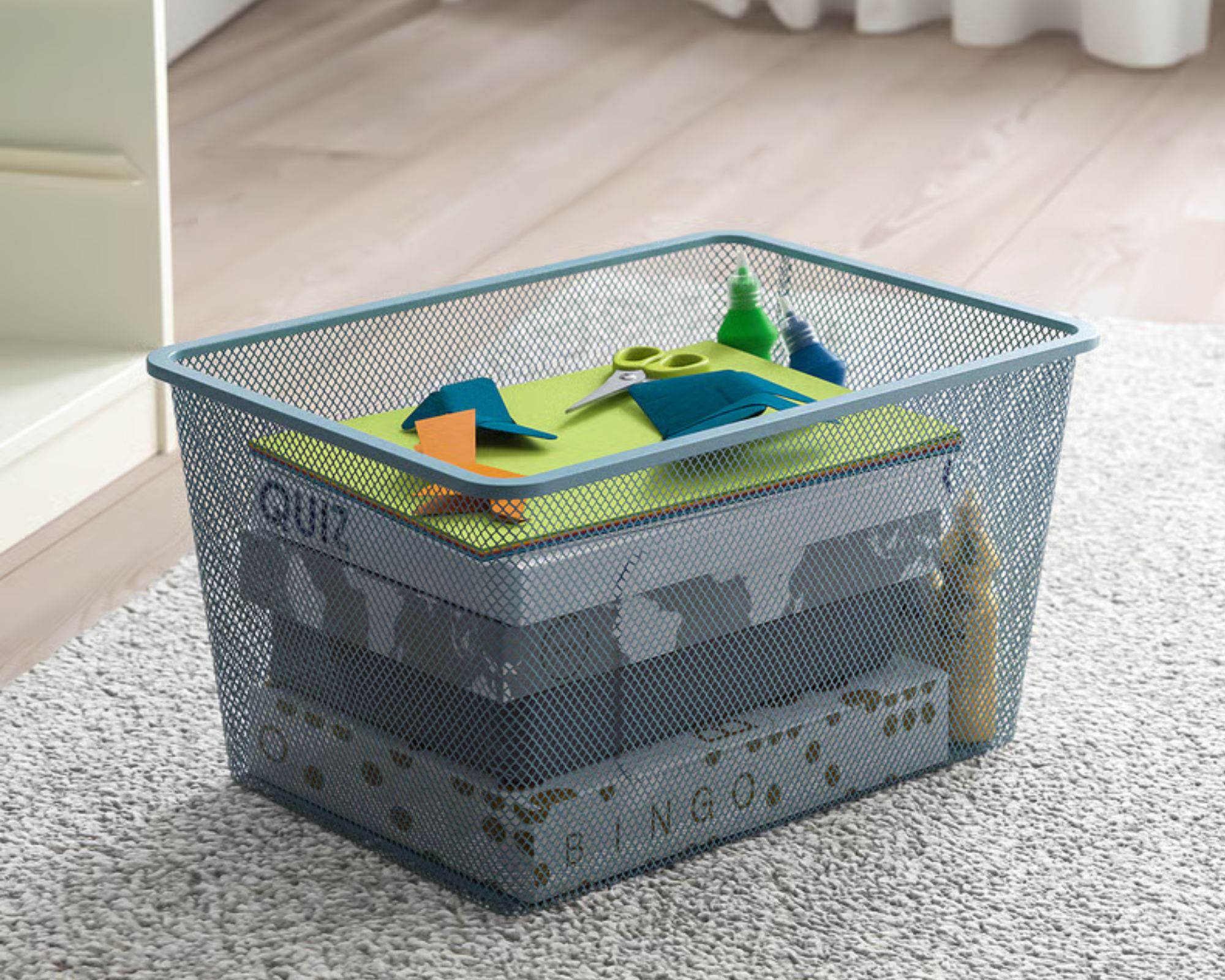 A pale blue IKEA TROFAST Mesh storage box filled with games, origami, and scissors, pictured on a wheat colored rug against light wood flooring.